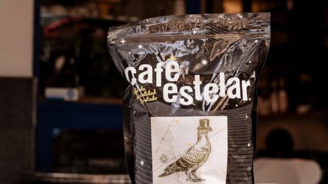 Paquete de café en grano de Café Estelar en una mesa, con taza y granos dispersos, en ambiente de ca.